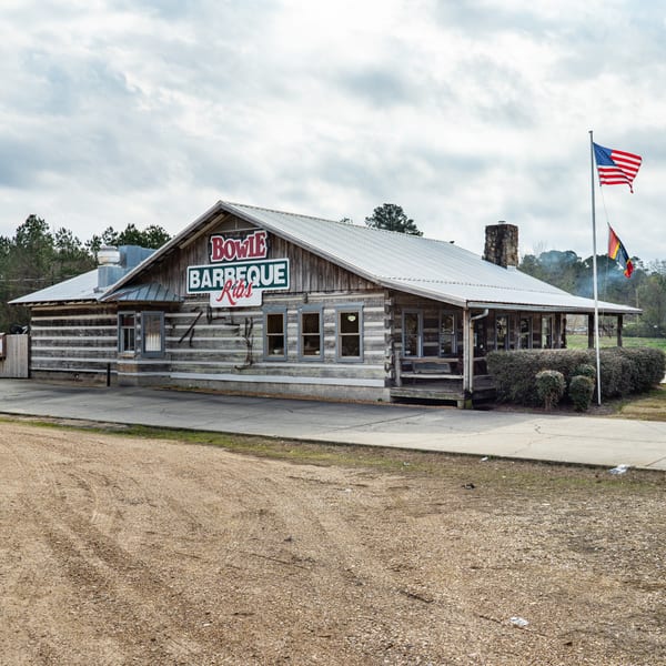 Exterior of Bowie Barbeque, a rustic log cabin building with a metal roof and American flag.